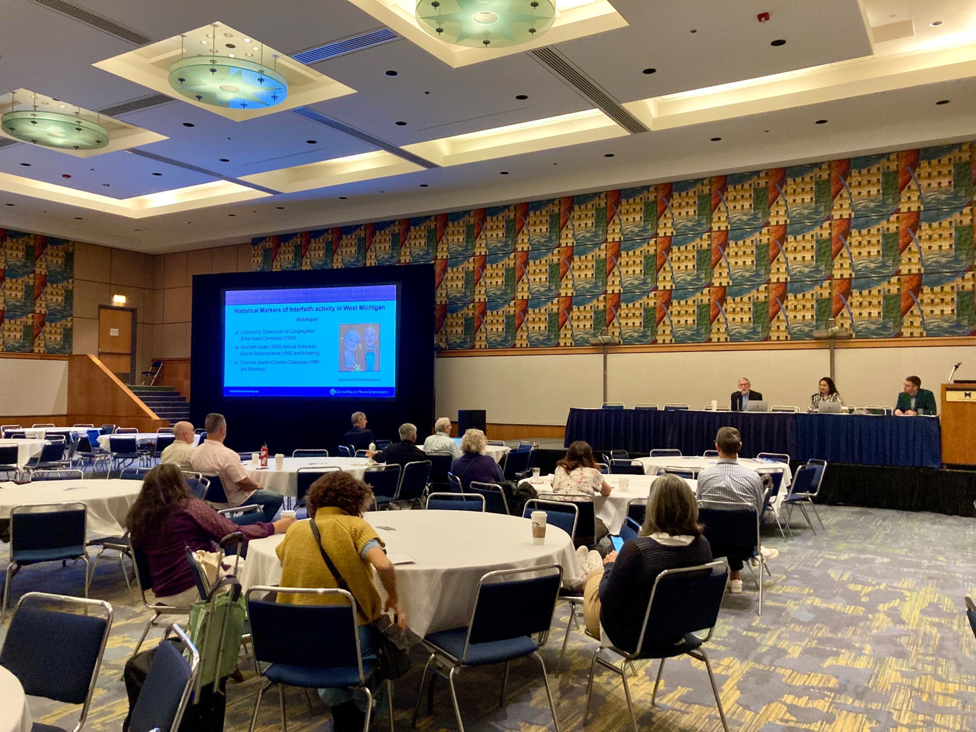 Three panelist and a presentation on screen in front of a room of people